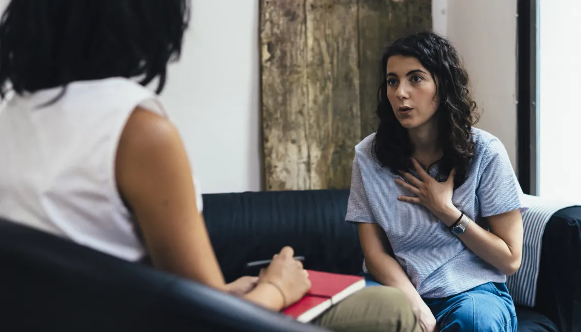 A woman talking across to another woman whilst sitting on a sofa.